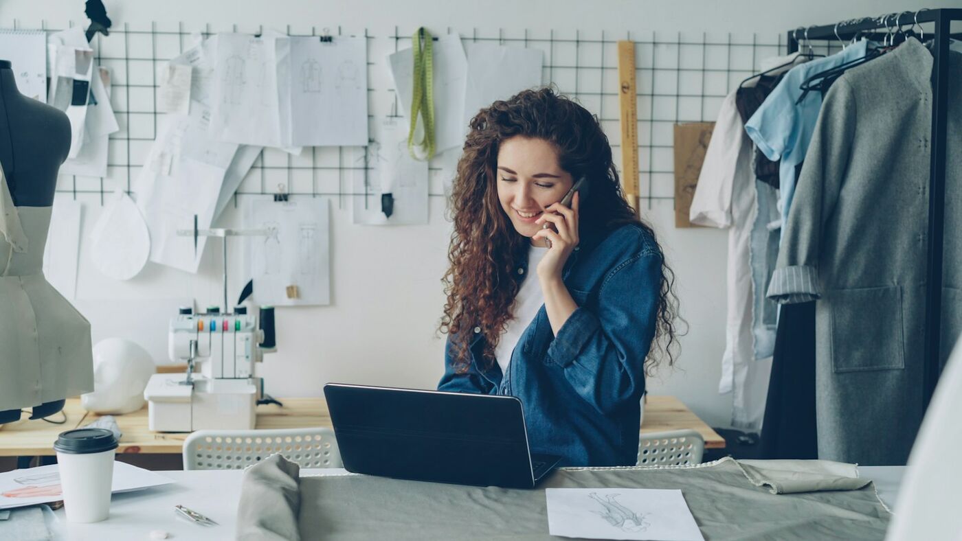 Small business owner taking a phone call while working on a laptop in her studio