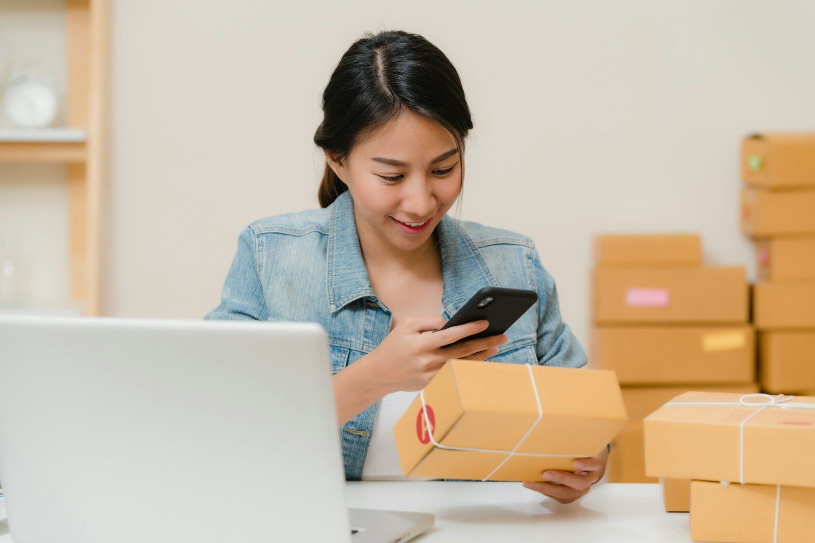 Small online store owner checking an order on her phone beside a laptop and stacked parcels